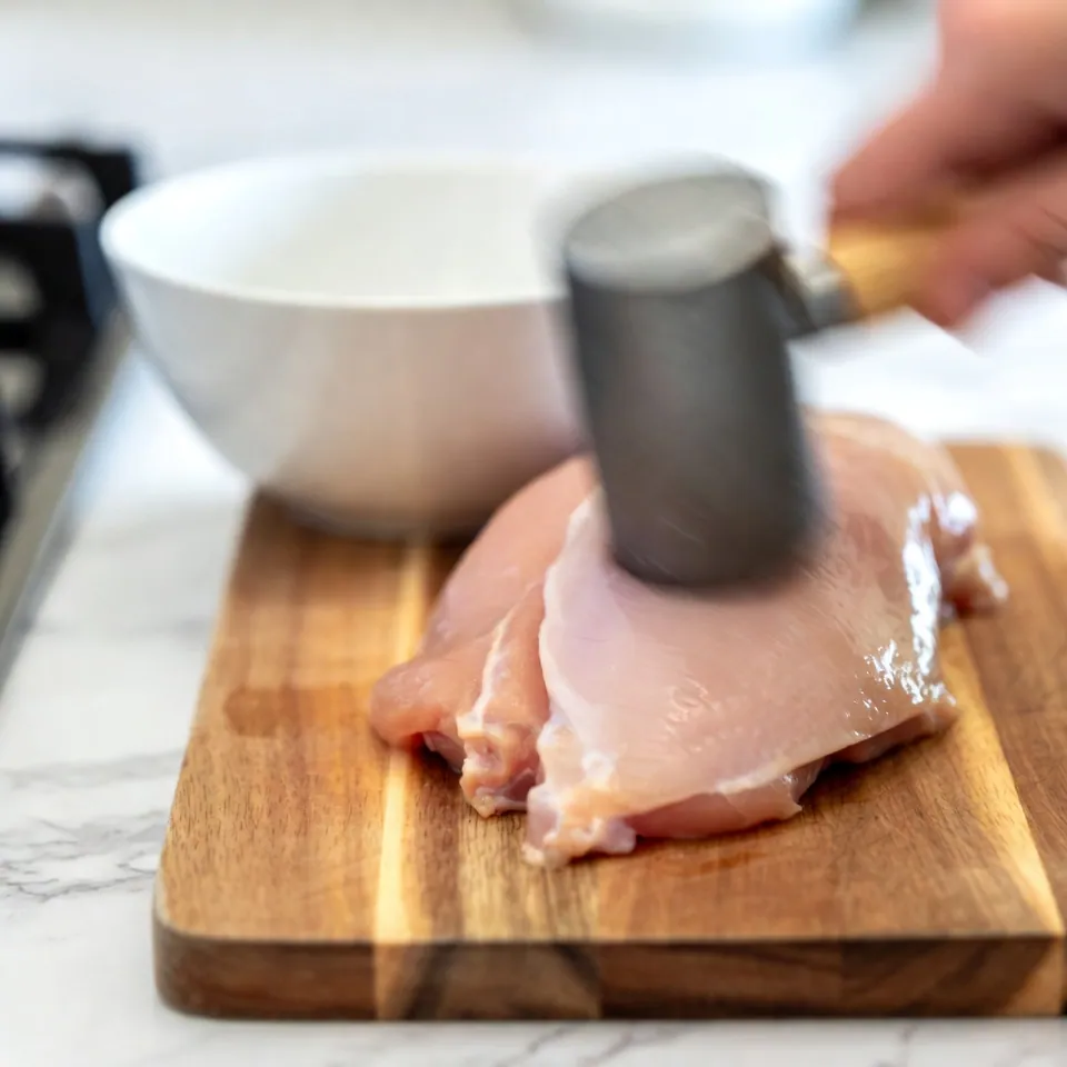 Chicken cutlets are being gently pounded with a meat mallet on a wooden cutting board